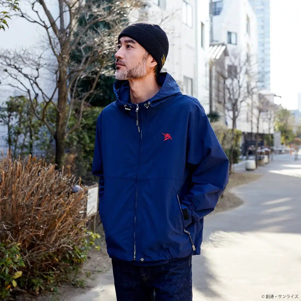 ビーニー帽をかぶり、ジオン軍のエンブレムが入った青いジャケットを着た男性が、屋外の道を歩いている様子。