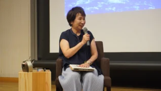 講演会かセミナーらしき場所で、笑顔でマイクを手に話しているアジア人女性の姿です。紺色のトップスとプリーツパンツを着用し、椅子に座っています。後方にはスクリーンが見えます。