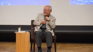 スーツジャケットを着た高齢の男性が椅子に座り、マイクを持って笑顔で話しています。講演会やイベントでの一幕のようです。