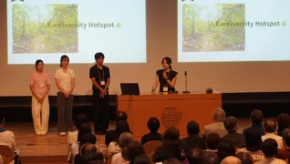 講堂のような会場でプレゼンテーションが行われている様子。壇上には4人の発表者がおり、そのうちの1人がマイクを持って話しています。背景のスクリーンには「A Biodiversity Hotspot」という文字と緑豊かな自然の画像が映し出されており、多数の聴衆が発表を聞いています。