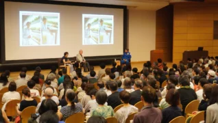 多くの聴衆が集まる会場で講演会が開催されており、壇上には3名の登壇者がいます。大型スクリーンには宇宙での植物工場実験に関する画像と文字が映し出されています。